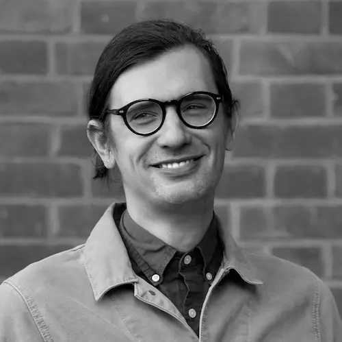 black-and-white photograph of elliot b. he is a 30-something man with fair skin, thick glasses, and dark brown hair. Behind him is a brick wall.